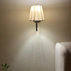 May include: A wall-mounted light fitting with a beige fabric lampshade. The lampshade has a pleated design and is attached to a silver metal arm.