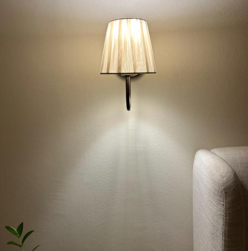 May include: A wall-mounted light fitting with a beige fabric lampshade. The lampshade has a pleated design and is attached to a silver metal arm.