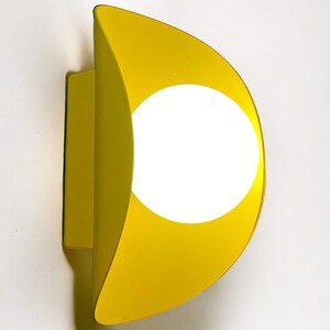 May include: A yellow wall sconce with a circular light fixture. The sconce is shaped like a crescent moon.