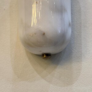 May include: A white marble-like object with a rounded bottom and a brass-colored metal base. The object is vertically oriented and appears to be a decorative item or a part of a larger fixture. The surface has subtle veining.