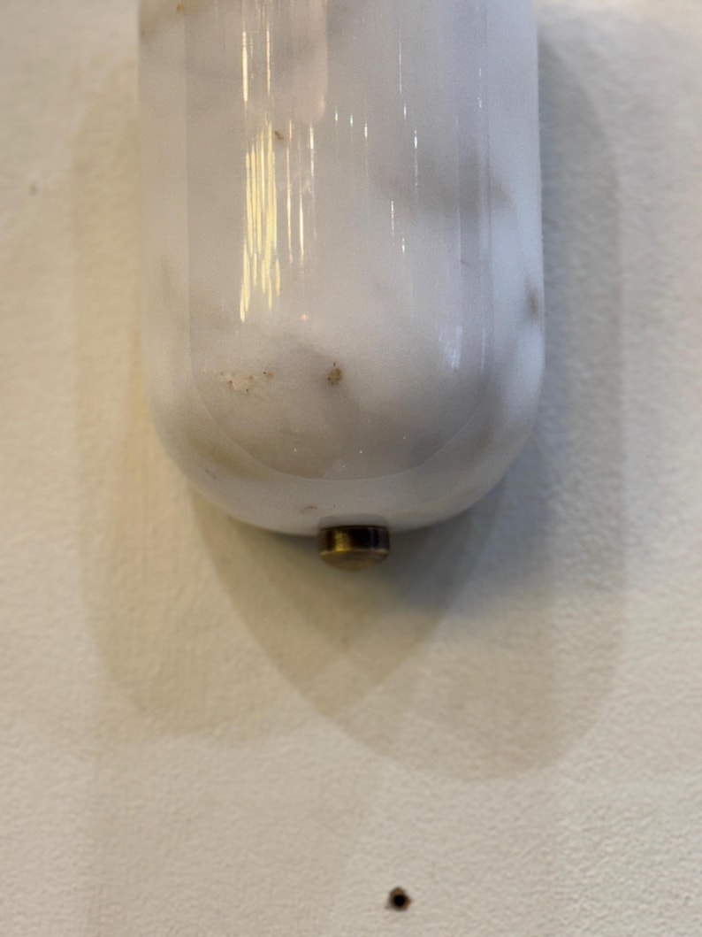May include: A white marble-like object with a rounded bottom and a brass-colored metal base. The object is vertically oriented and appears to be a decorative item or a part of a larger fixture. The surface has subtle veining.