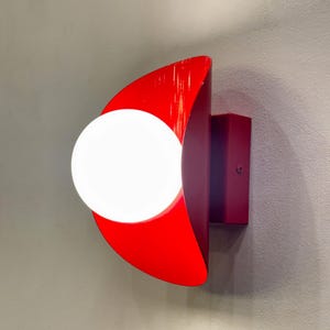 May include: A red and burgundy wall sconce with a white globe light bulb. The sconce is shaped like a crescent moon.