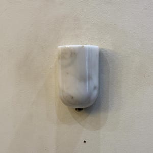May include: A white marble object with gray veining, possibly a decorative piece or furniture component. The object has a rounded bottom and a small metal fitting. The background is a plain, off-white surface.