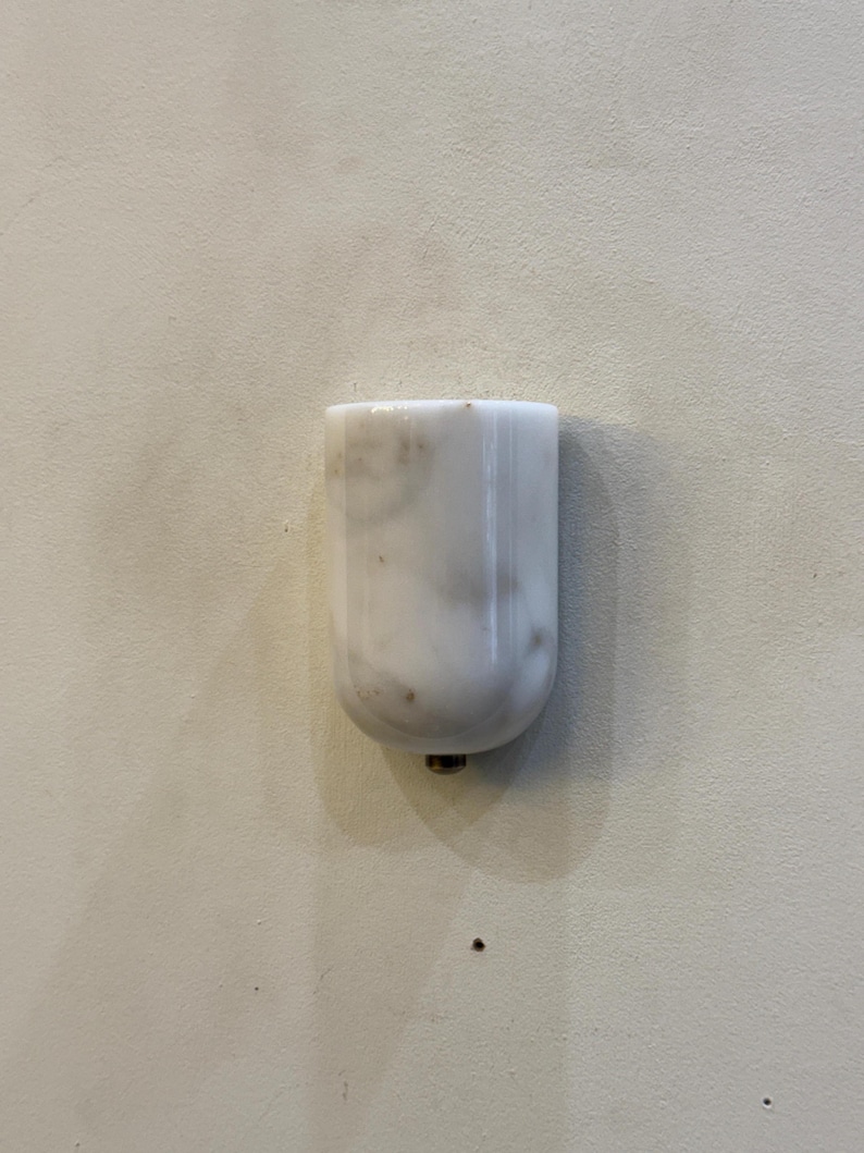 May include: A white marble object with gray veining, possibly a decorative piece or furniture component. The object has a rounded bottom and a small metal fitting. The background is a plain, off-white surface.