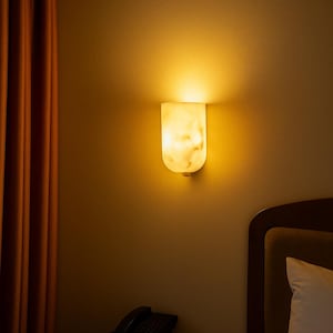 May include: A wall-mounted, illuminated sconce with a marble-like appearance. The light fixture emits a warm, inviting glow. The sconce is mounted on a beige wall, with a dark brown nightstand, a telephone, and a remote control visible below.