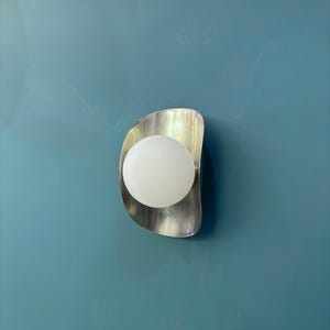 May include: A contemporary wall light featuring a brushed metal surround and a spherical, frosted glass bulb. The light is mounted on a teal wall, offering a minimalist design. The light fixture is designed for indoor use.