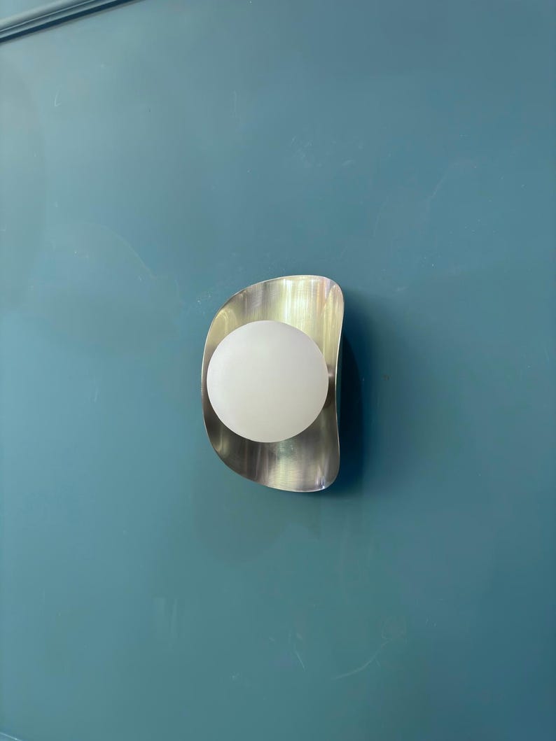 May include: A contemporary wall light featuring a brushed metal surround and a spherical, frosted glass bulb. The light is mounted on a teal wall, offering a minimalist design. The light fixture is designed for indoor use.