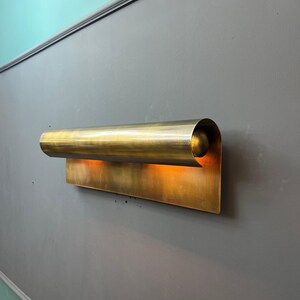May include: A brass wall sconce with a curved, cylindrical top and a rectangular base. The sconce emits a warm, orange glow, creating a soft, ambient light. The fixture is mounted on a gray wall.
