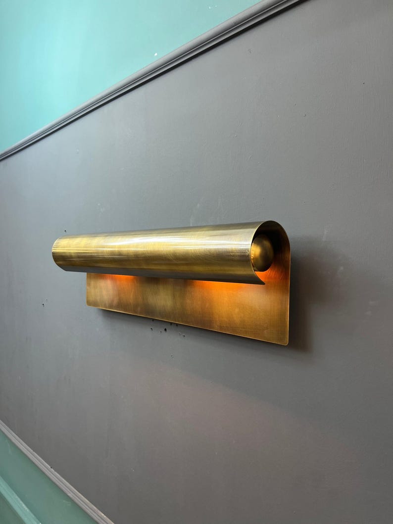 May include: A brass wall sconce with a curved, cylindrical top and a rectangular base. The sconce emits a warm, orange glow, creating a soft, ambient light. The fixture is mounted on a gray wall.