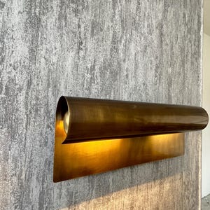 May include: A modern, brass-toned wall sconce with a curved, cylindrical shade and a rectangular base. The sconce is mounted on a textured, gray wall, and the light from the fixture illuminates the wall below.