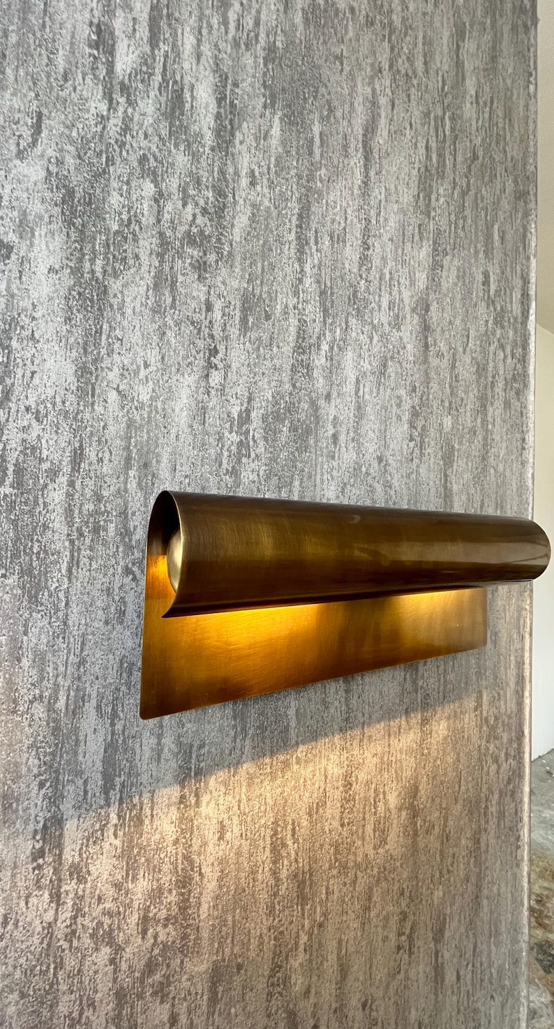 May include: A modern, brass-toned wall sconce with a curved, cylindrical shade and a rectangular base. The sconce is mounted on a textured, gray wall, and the light from the fixture illuminates the wall below.