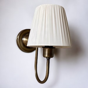 May include: A brass wall sconce with a white fabric lampshade. The sconce has a curved arm and a round base.
