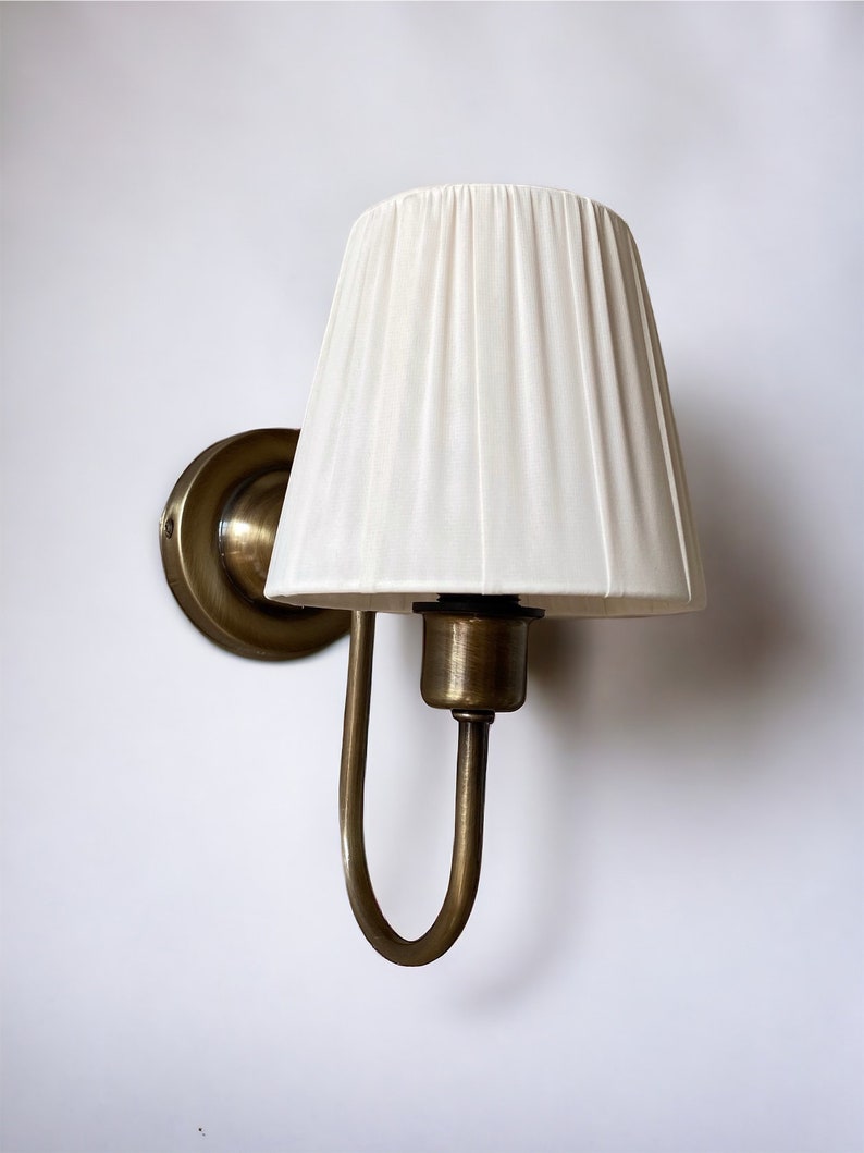 May include: A brass wall sconce with a white fabric lampshade. The sconce has a curved arm and a round base.