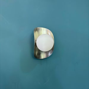 May include: A contemporary wall light featuring a brushed metal backplate and a spherical, frosted glass bulb. The sconce is mounted on a teal wall, offering a contrast of textures and colours. The design is minimalist and modern.