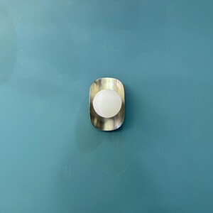 May include: A contemporary wall light with a brushed metal surround and a spherical, frosted glass bulb. The light is fixed to a teal wall with decorative molding.