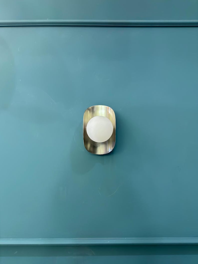 May include: A contemporary wall light with a brushed metal surround and a spherical, frosted glass bulb. The light is fixed to a teal wall with decorative molding.