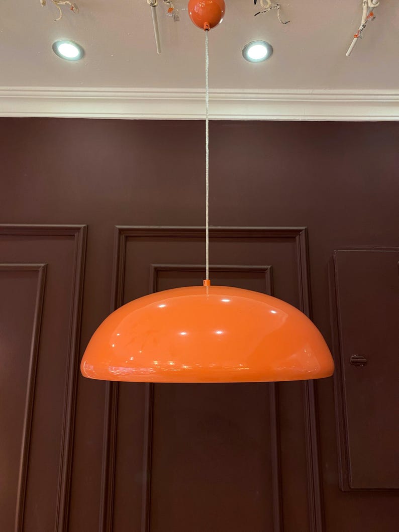 May include: A bright orange, dome-shaped pendant light fixture hangs from a white cord against a dark brown wall.