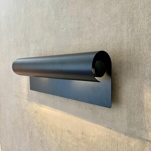 May include: A modern, black wall sconce with a cylindrical shade and a rectangular base. The sconce is made of metal and emits a warm light. The design is minimalist and contemporary, suitable for indoor or outdoor use.