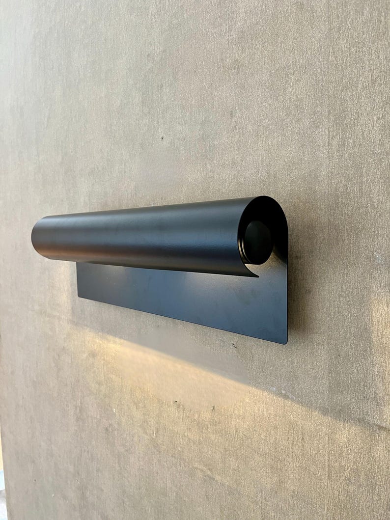 May include: A modern, black wall sconce with a cylindrical shade and a rectangular base. The sconce is made of metal and emits a warm light. The design is minimalist and contemporary, suitable for indoor or outdoor use.