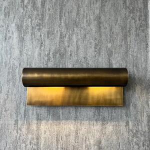 May include: A modern wall sconce with a cylindrical top and a rectangular base, both in a warm brass finish. The sconce emits a soft, golden light, creating a welcoming ambiance. The fixture is mounted on a textured gray wall.