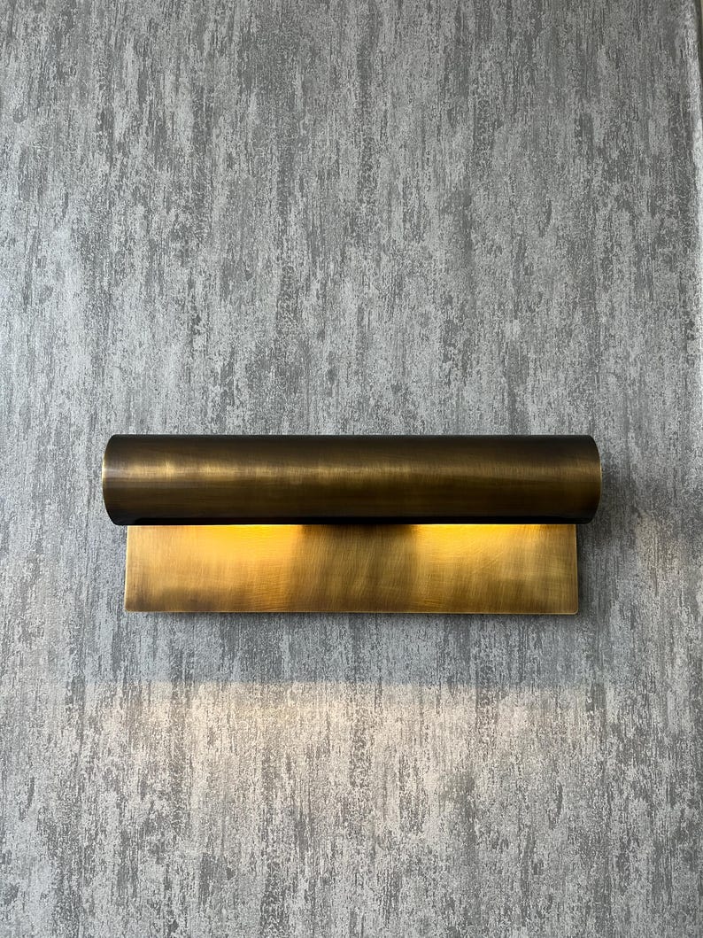 May include: A modern wall sconce with a cylindrical top and a rectangular base, both in a warm brass finish. The sconce emits a soft, golden light, creating a welcoming ambiance. The fixture is mounted on a textured gray wall.