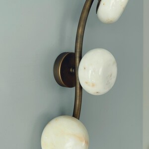 May include: A wall sconce with a curved brass arm and three white, spherical shades. The shades have a marbled appearance. The sconce is mounted on a round brass base, creating a modern lighting fixture.