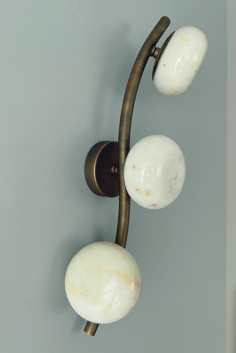 May include: A wall sconce with a curved brass arm and three white, spherical shades. The shades have a marbled appearance. The sconce is mounted on a round brass base, creating a modern lighting fixture.