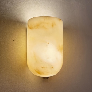 May include: A wall-mounted, cylindrical sconce with a warm, glowing light. The fixture is made of a translucent, creamy-yellow material with subtle brown veining. The sconce is mounted on a neutral-colored wall, casting a soft shadow.