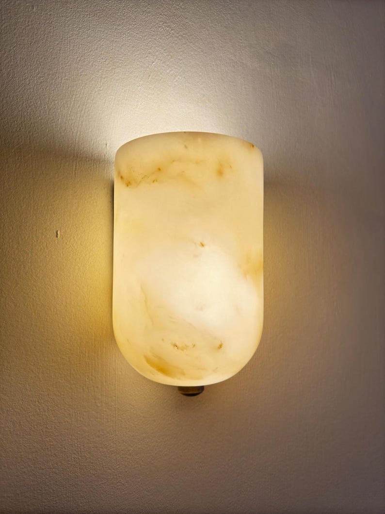 May include: A wall-mounted, cylindrical sconce with a warm, glowing light. The fixture is made of a translucent, creamy-yellow material with subtle brown veining. The sconce is mounted on a neutral-colored wall, casting a soft shadow.