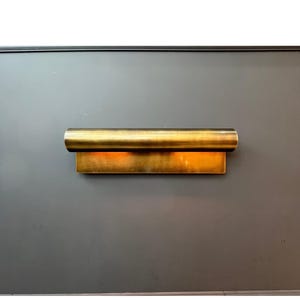 May include: A modern, gold-toned wall sconce. The fixture has a rectangular base with a cylindrical top, emitting a warm, inviting glow. The sconce is mounted on a dark gray wall, creating a striking contrast.
