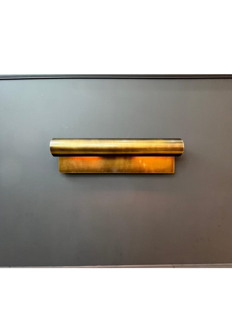 May include: A modern, gold-toned wall sconce. The fixture has a rectangular base with a cylindrical top, emitting a warm, inviting glow. The sconce is mounted on a dark gray wall, creating a striking contrast.