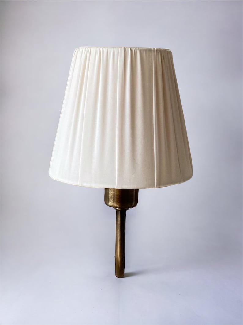 May include: A white pleated lampshade with a brass wall sconce.