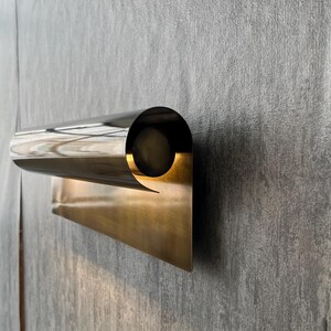 May include: A modern wall sconce with a polished, metallic finish. The fixture features a cylindrical light housing and a rectangular base, creating a minimalist design. The sconce is mounted on a textured gray wall.