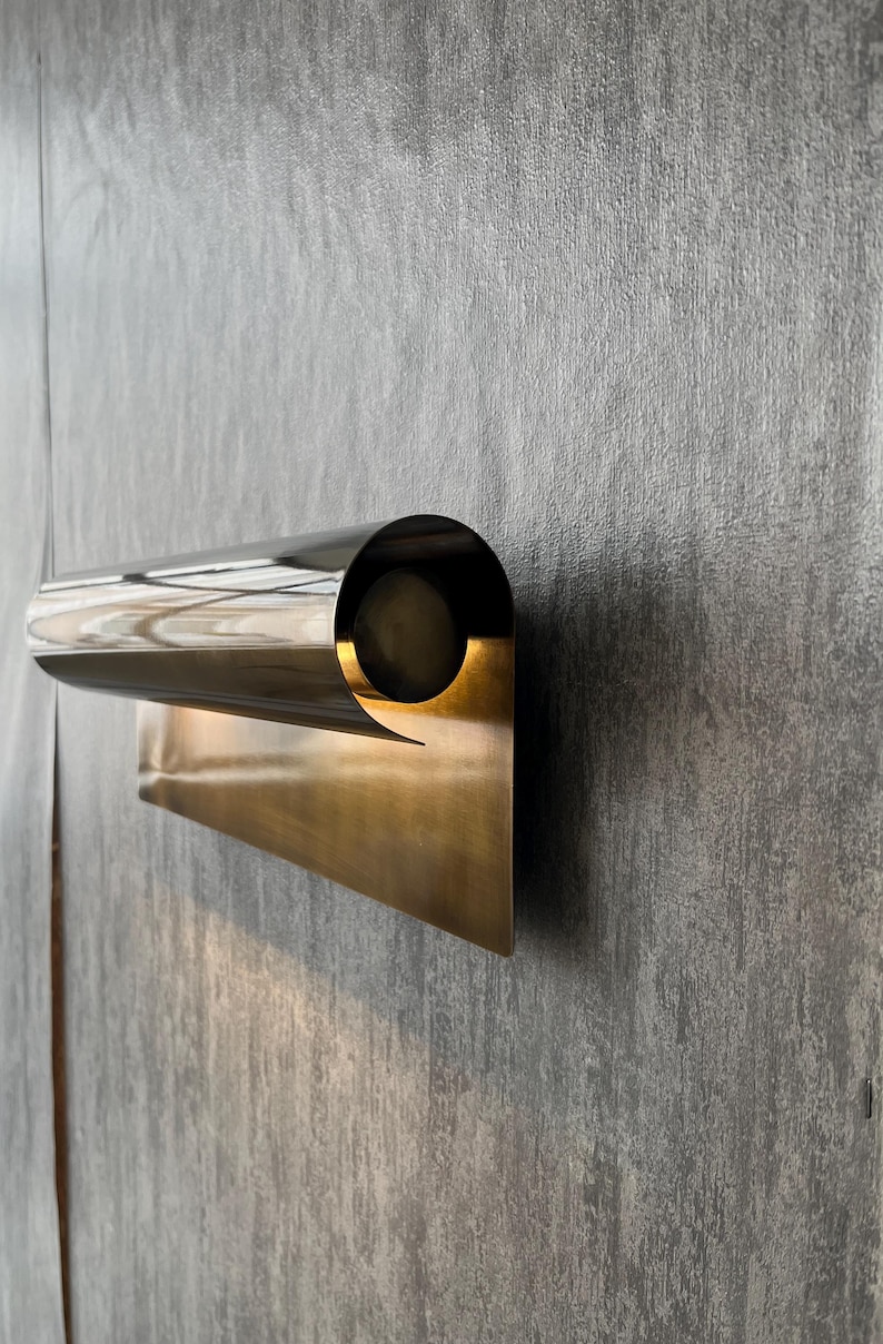 May include: A modern wall sconce with a polished, metallic finish. The fixture features a cylindrical light housing and a rectangular base, creating a minimalist design. The sconce is mounted on a textured gray wall.
