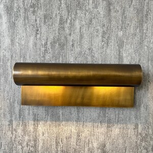 May include: A brass wall sconce with a cylindrical top and a rectangular base. The sconce emits a warm, golden light, creating a soft glow. The fixture is mounted on a textured, gray wall.