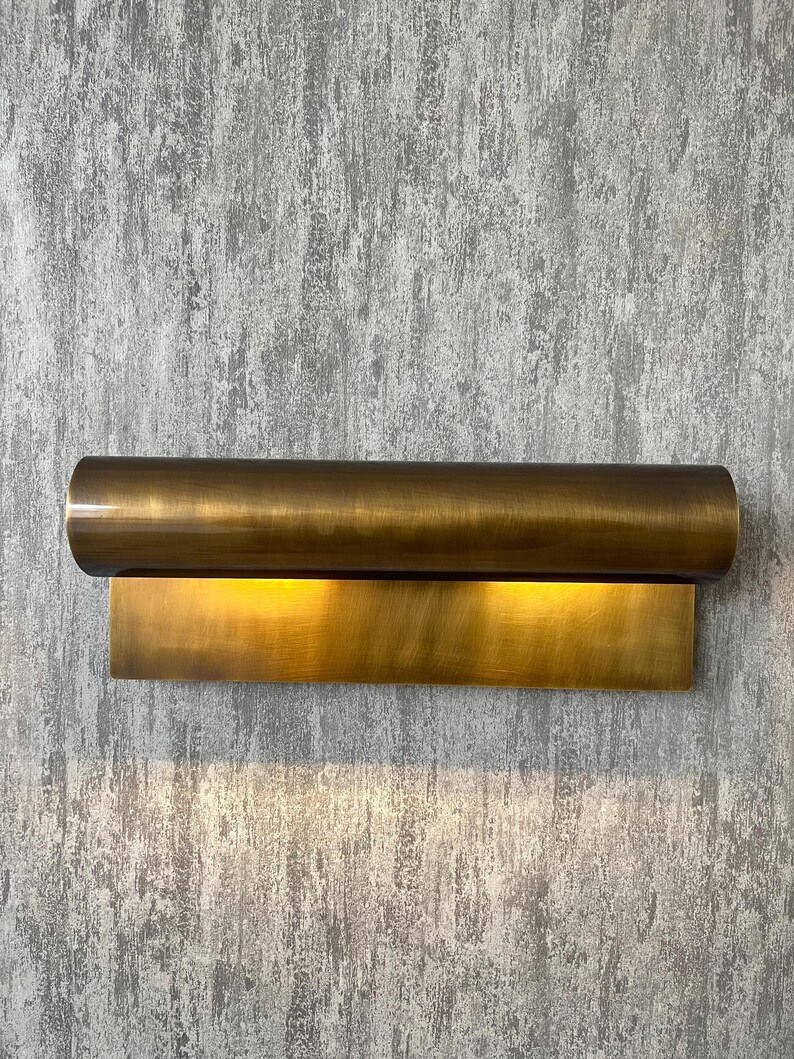 May include: A brass wall sconce with a cylindrical top and a rectangular base. The sconce emits a warm, golden light, creating a soft glow. The fixture is mounted on a textured, gray wall.