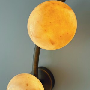 May include: A wall-mounted light fixture featuring two spherical, glowing orbs. The orbs have a warm, yellow-orange hue with subtle variations in color. The fixture's arm and base are a dark, bronze-like color, creating a contrast with the light orbs.