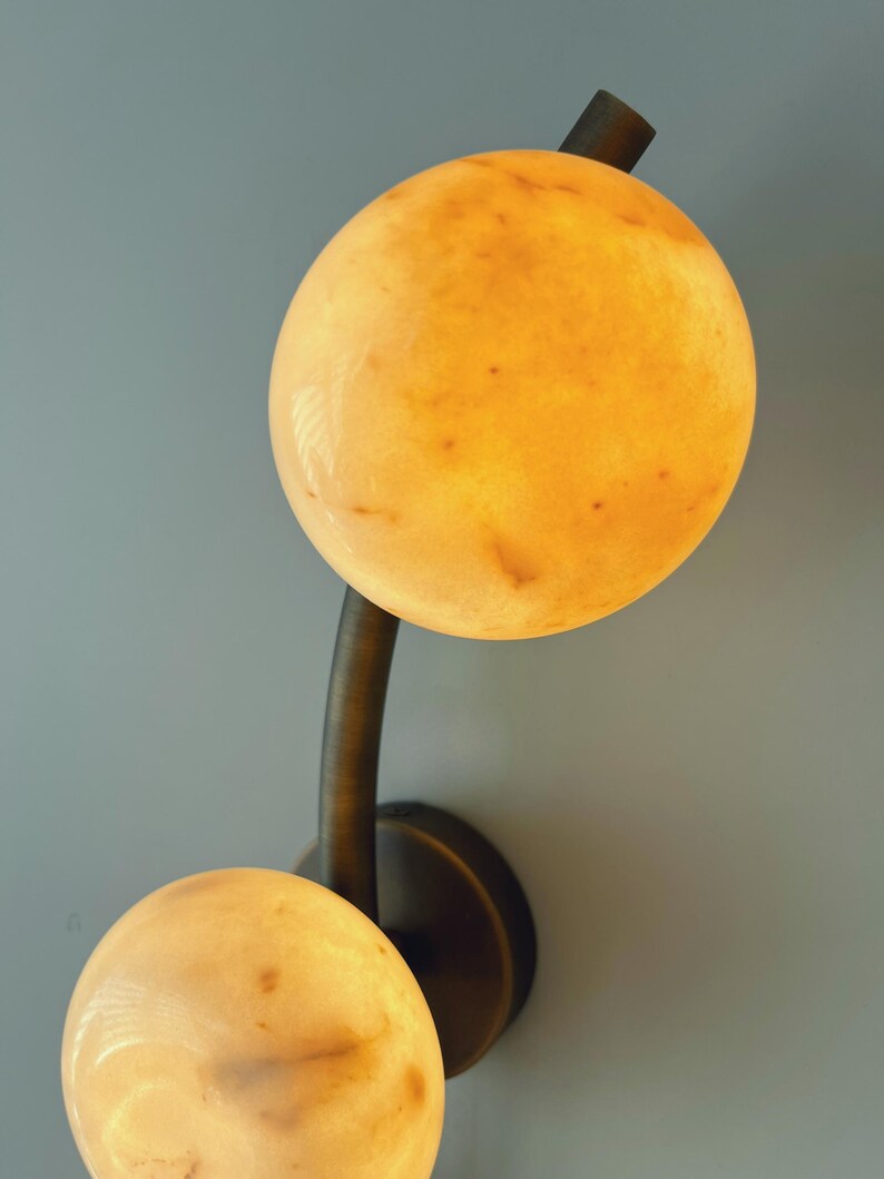 May include: A wall-mounted light fixture featuring two spherical, glowing orbs. The orbs have a warm, yellow-orange hue with subtle variations in color. The fixture's arm and base are a dark, bronze-like color, creating a contrast with the light orbs.