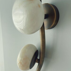 May include: A wall-mounted light fixture with two white, marbled glass globes. The globes are attached to a curved, bronze-colored metal arm and a circular base. The fixture is designed for indoor use and adds a touch of elegance.