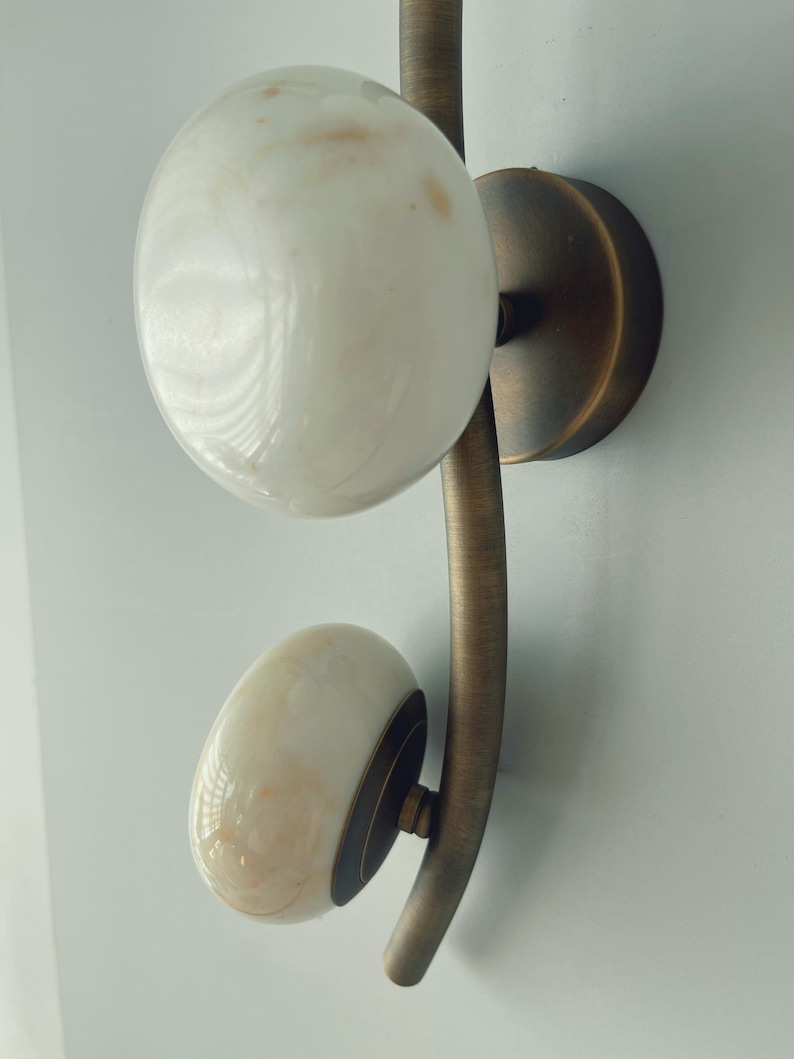 May include: A wall-mounted light fixture with two white, marbled glass globes. The globes are attached to a curved, bronze-colored metal arm and a circular base. The fixture is designed for indoor use and adds a touch of elegance.