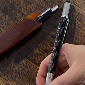 May include: A black multi-functional pen featuring a silver knurled grip, a built-in ruler with markings from 0 to 3 inches, and a stylus tip. A second pen with the name Lombardo printed on the side rests inside a brown leather sleeve.