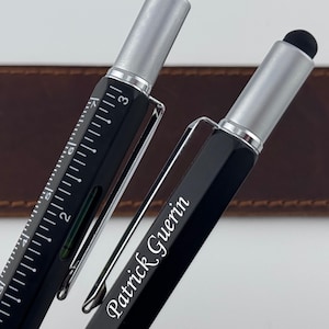 May include: Two black multi-functional pens with silver accents and black stylus tips. One pen features a white printed ruler with inch markings and a small level. The other pen is personalized with the white text Patrick Guerin.