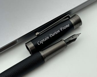 May include: A black fountain pen with a silver trim and a black and silver cap. The cap has the inscription "Captain Darian Frame" in white lettering.