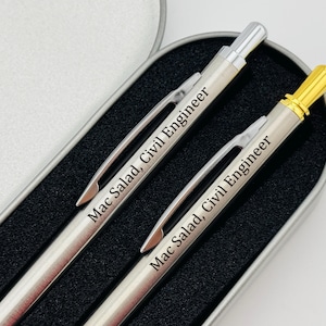 May include: Two silver pens with gold accents in a black and silver case. The pens are engraved with the text "Mac Salad, Civil Engineer".