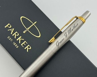 May include: A silver-toned metal ballpoint pen with a gold-colored clip and the engraved name Grace J Mathes, resting on a black Parker gift box featuring the gold Parker logo and the text PARKER EST. 1888 printed on the surface of the box.