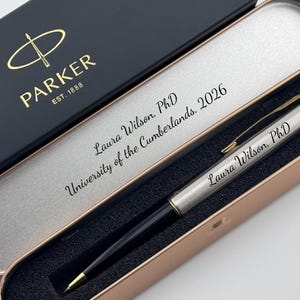 May include: A silver and black Parker pen with gold accents, presented in a black box with the Parker logo. The pen is engraved with "Laura Wilson, PhD" and the box reads "PARKER EST. 1888". The pen case is open, revealing the pen and the inscription "University of the Cumberlands, 2026".
