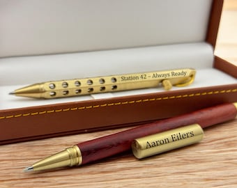 May include: Two pens in a brown leatherette box. One pen is gold-colored with the text "Station 42 - Always Ready." The other pen is red and gold with the text "Aaron Eilers" on the cap. The pens are on a wooden surface.
