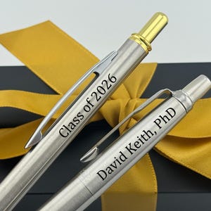 May include: Two silver pens with black text, "Class of 2026" and "David Keith, PhD." The pens have gold accents and silver clips. A gold ribbon and a black box are in the background.