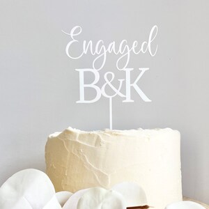 May include: White cake topper with the words "Engaged" and "B&K" in a cursive font. The topper is on top of a white cake.