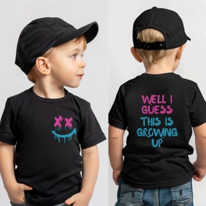 May include: A black t-shirt featuring a pink and blue dripping smiley face graphic on the front chest. The back of the shirt displays the text "WELL I GUESS THIS IS GROWING UP" in pink and blue lettering. The shirt is paired with a black baseball cap.